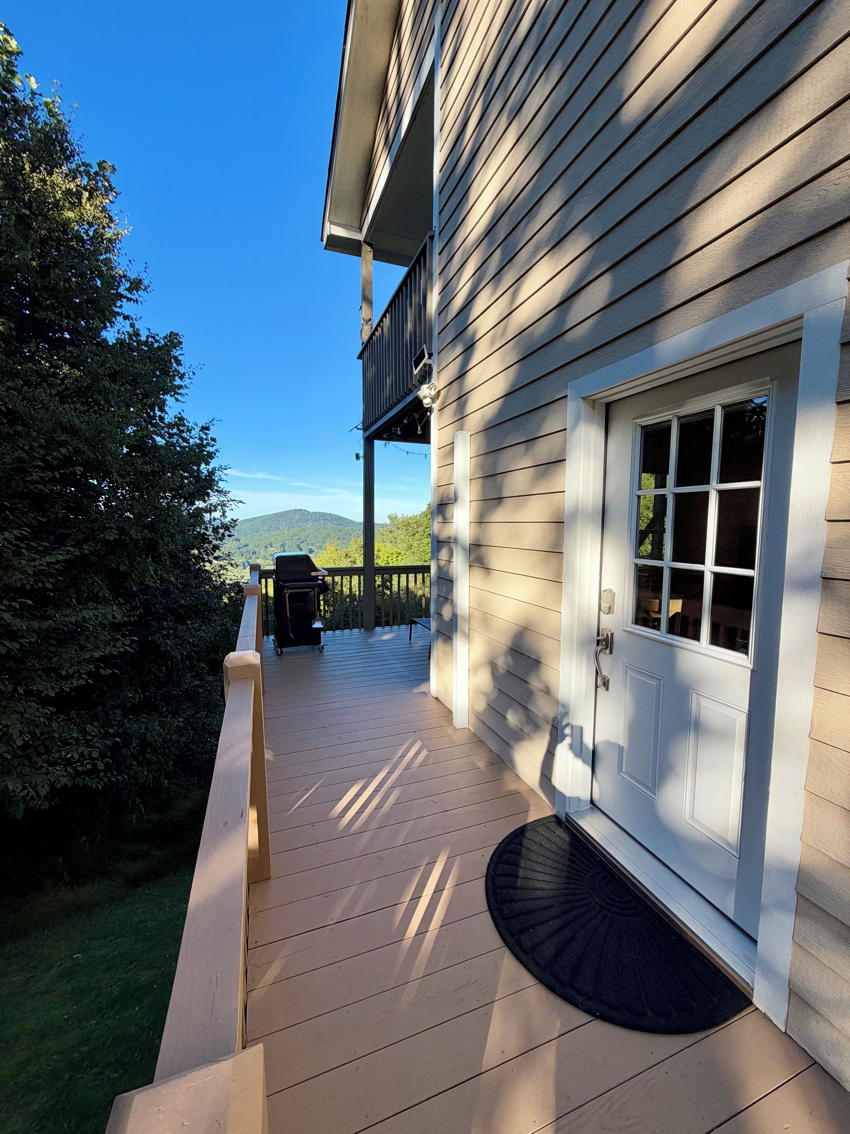 Une terrasse en bois avec une porte et une fenêtre, entourée de verdure et sous un ciel dégagé.