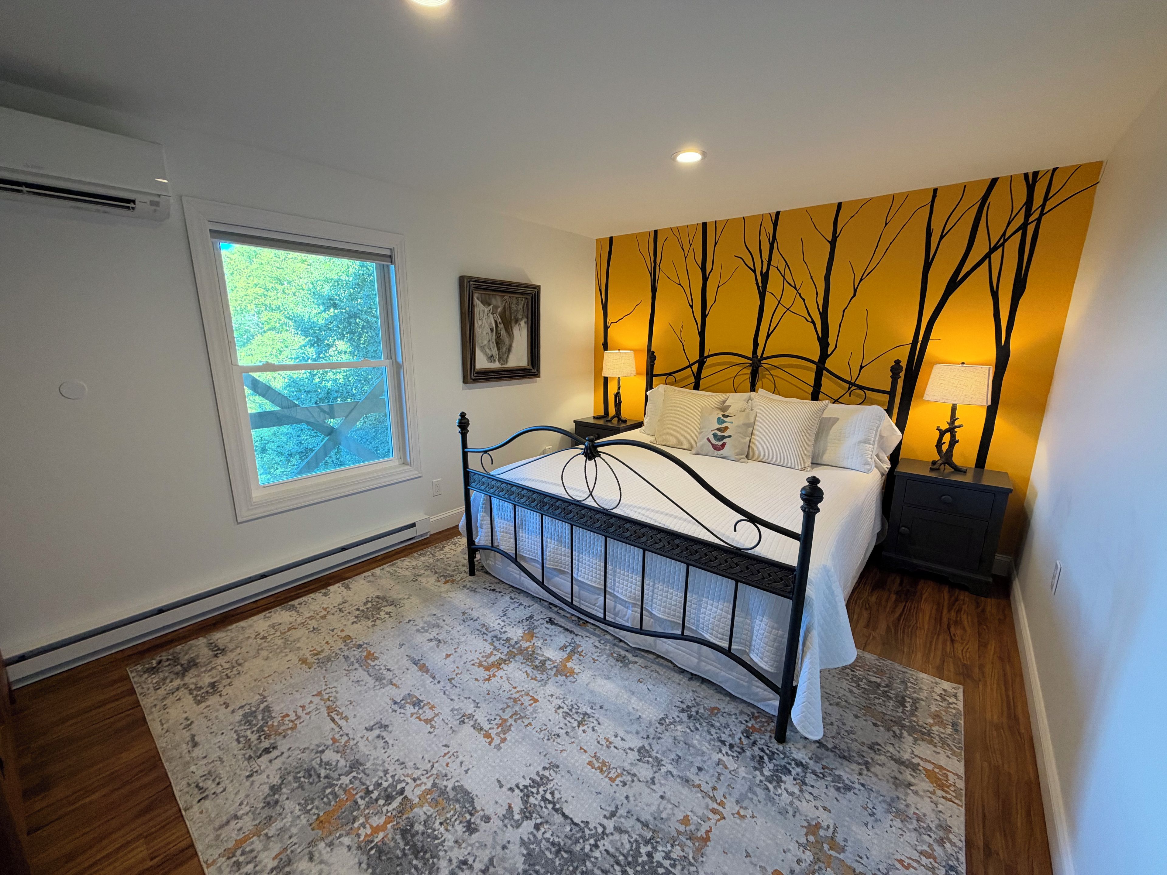A bedroom with a bed, a window, a wall with a tree mural, and a nightstand with a lamp.