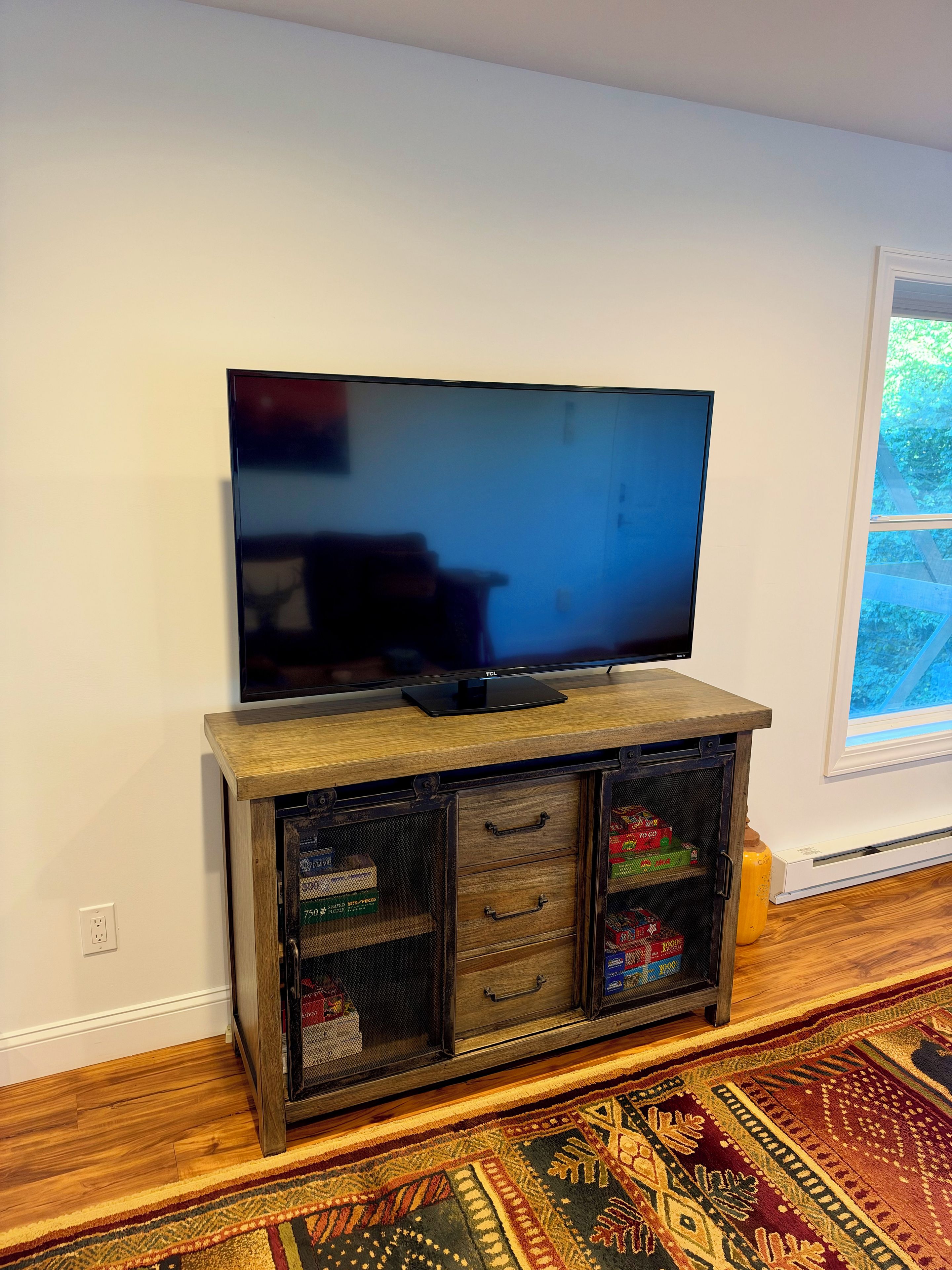 A flat-screen TV on a wooden cabinet with drawers and shelves, a patterned rug, and a window with a view of greenery.