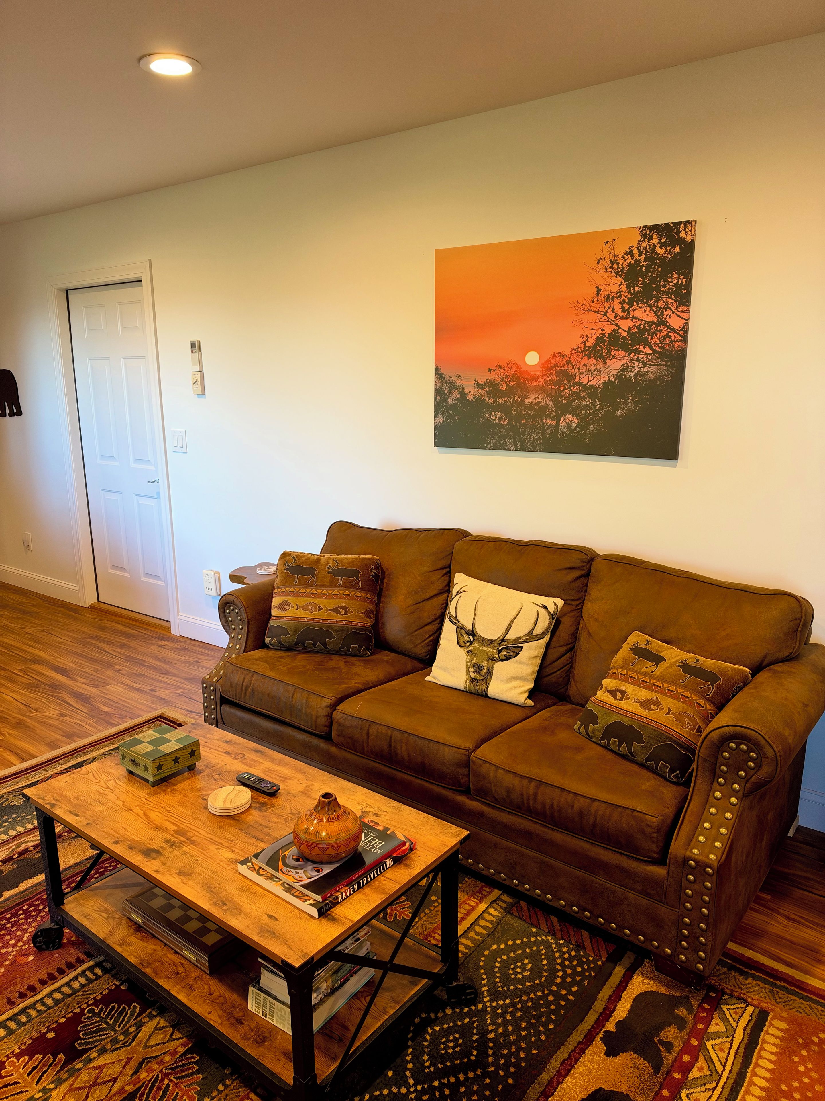 A living room with a brown leather sofa, a wooden coffee table with books and a globe, a patterned rug, and a sunset painting on the wall.