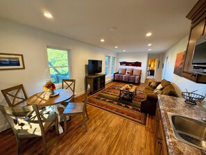 A modern living room with a dining area, wooden flooring, a flat-screen TV, and a kitchen with a built-in oven. - Serene Sunrise Long Range Mountain View 1-bedroom home in charming Blowing Rock (Blowing Rock)