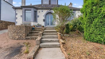 A two-story house with a stone staircase leading to the front door, surrounded by greenery and a brick pathway.