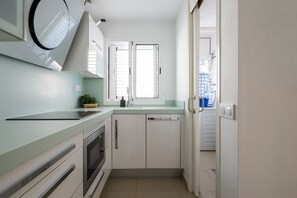A compact kitchen with a microwave, oven, and sink. There is a window with blinds, a door leading to a laundry area, and a small plant on the counter. - DA CESAR | Spacious Stay near Maspalomas (El Tablero)