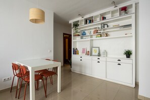Un coin repas avec une table blanche et des chaises orange, un meuble blanc avec des étagères et un luminaire suspendu.