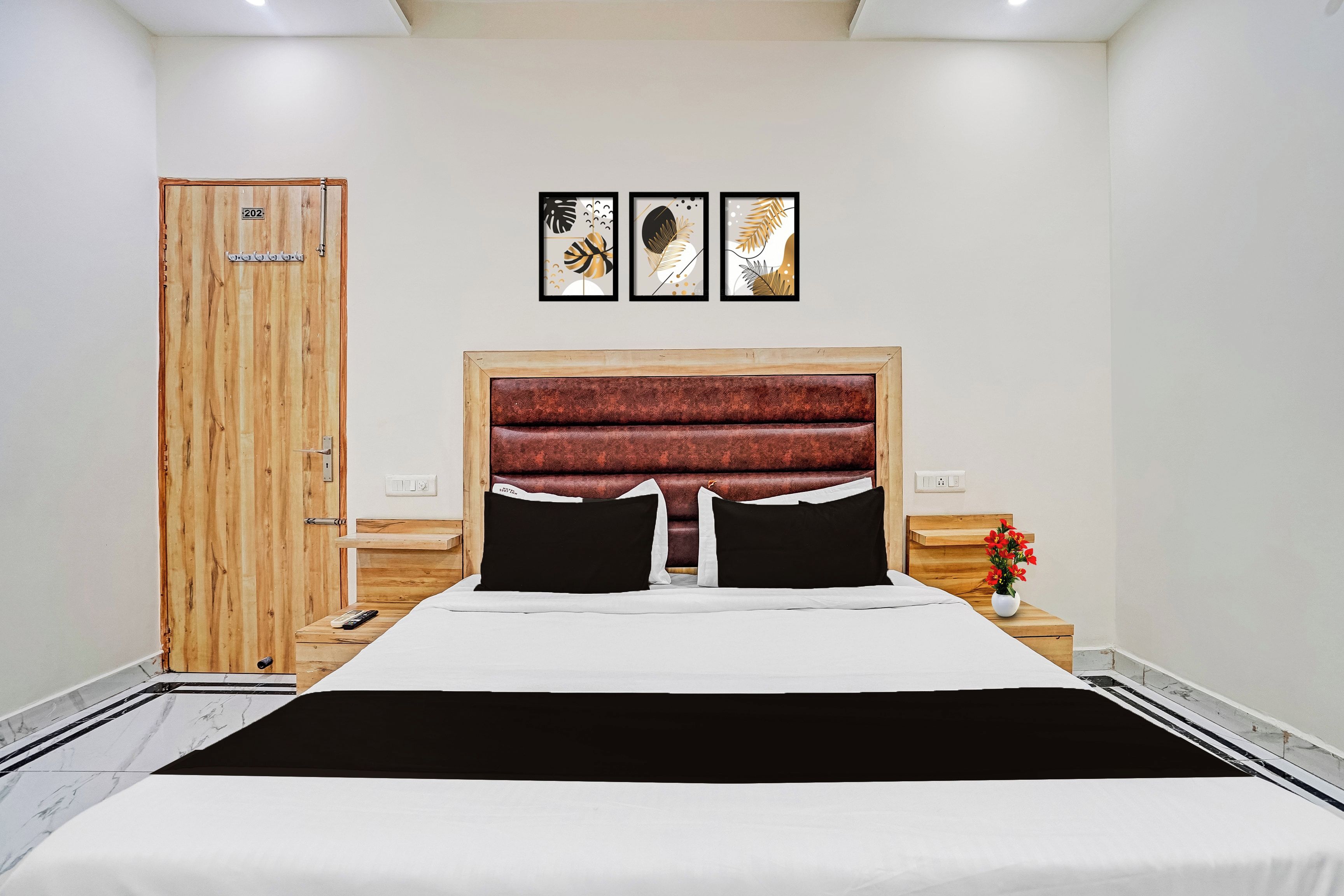 Habitación de hotel con puerta de madera, cama con cabecera roja, mesita de noche con un jarrón de flores y tres obras de arte enmarcadas en la pared.