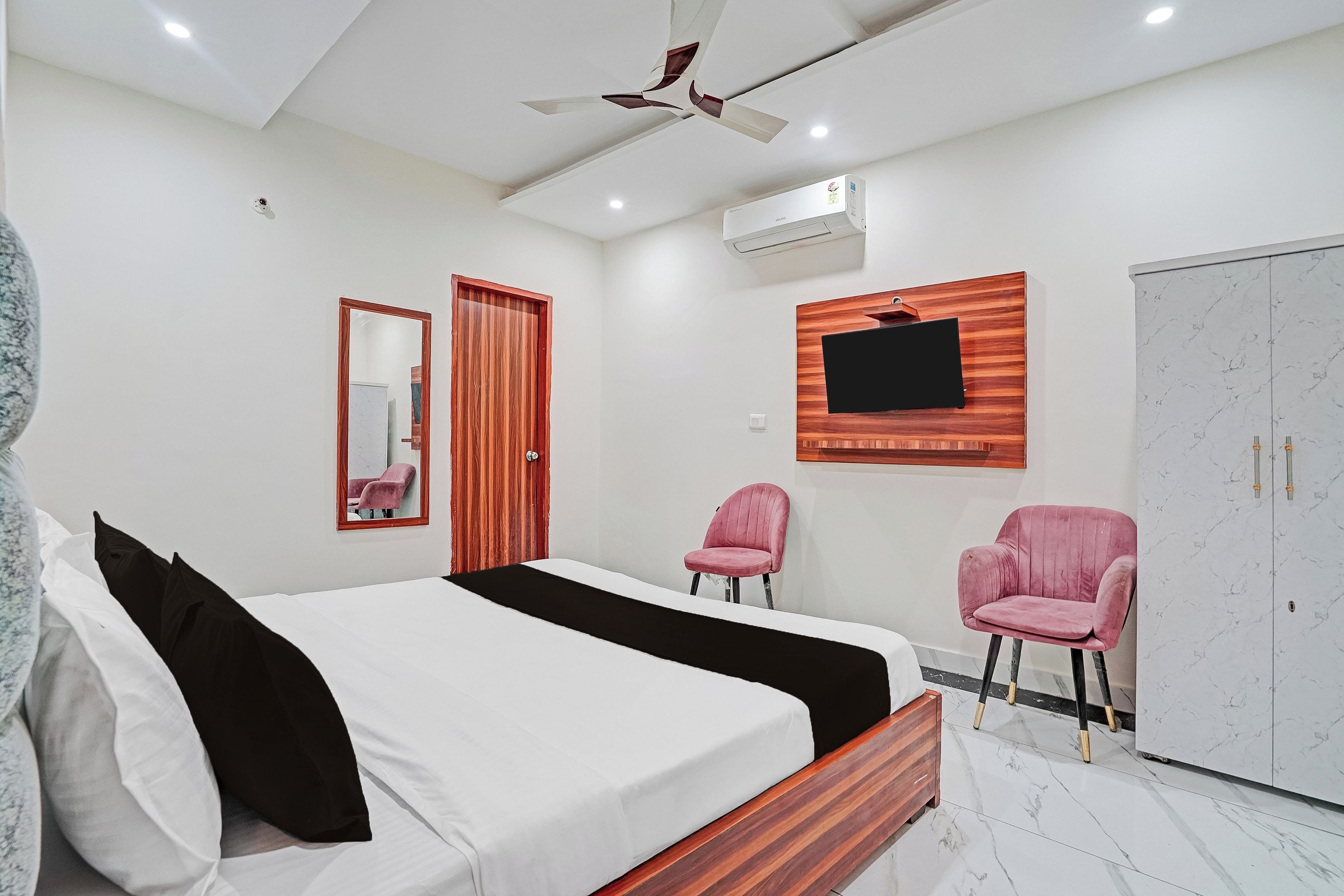 A hotel room with a bed, two chairs, a wardrobe, a ceiling fan, and an air conditioning unit.