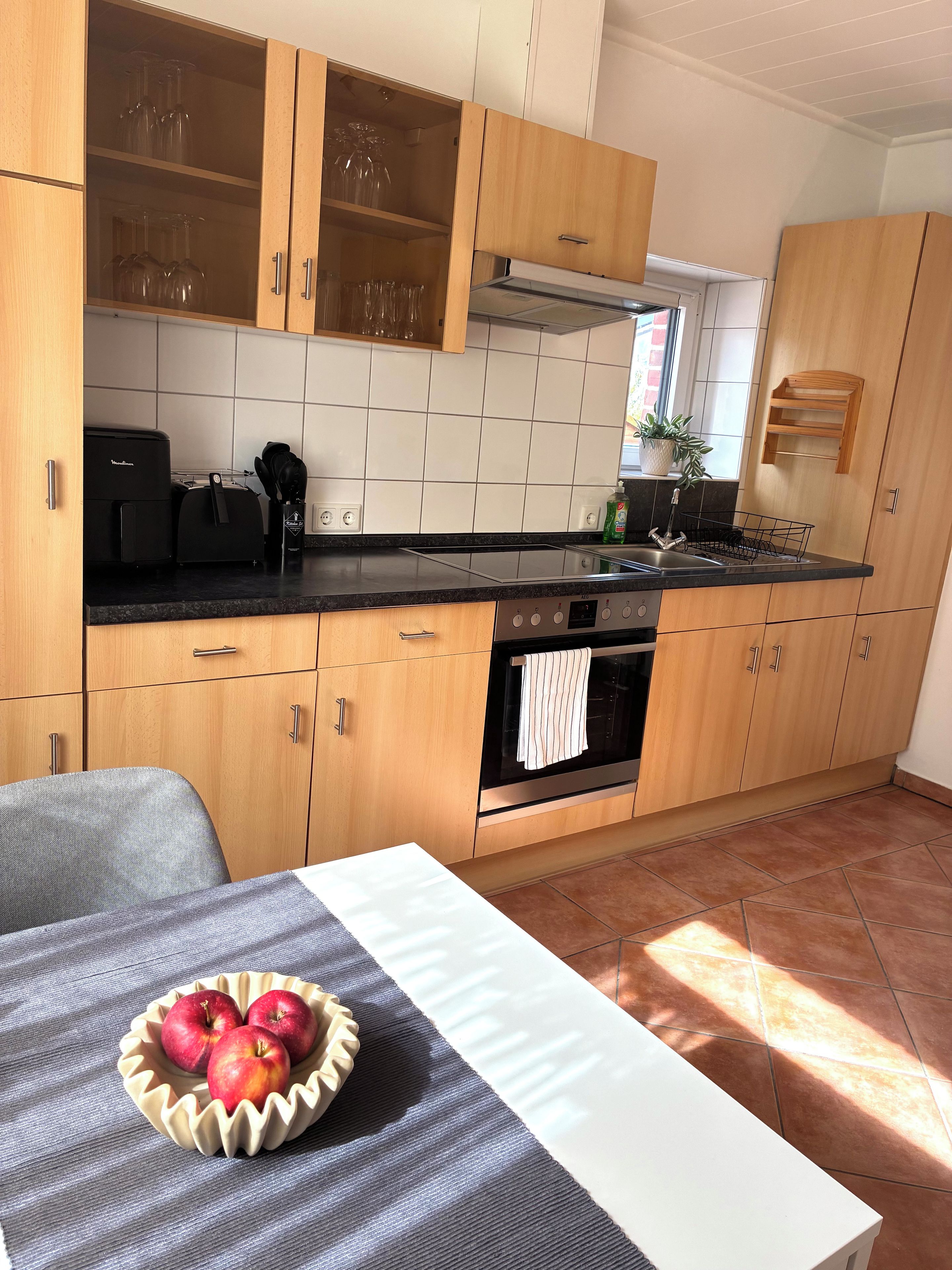 A kitchen with wooden cabinets, a black countertop, a built-in oven, and a dining area with a table and a bowl of fruit.