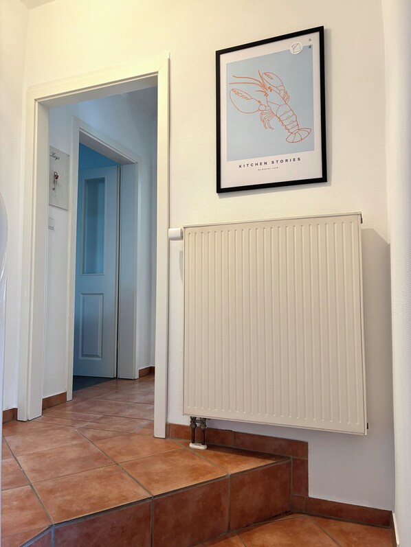 A hallway with a framed artwork above a radiator, a door with a nameplate, and a tiled floor.