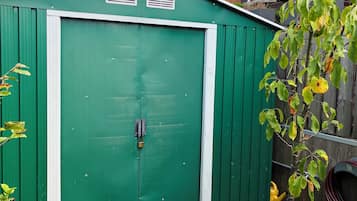 Un cobertizo metálico verde con una puerta blanca, un árbol pequeño en una maceta y una valla de madera al fondo.