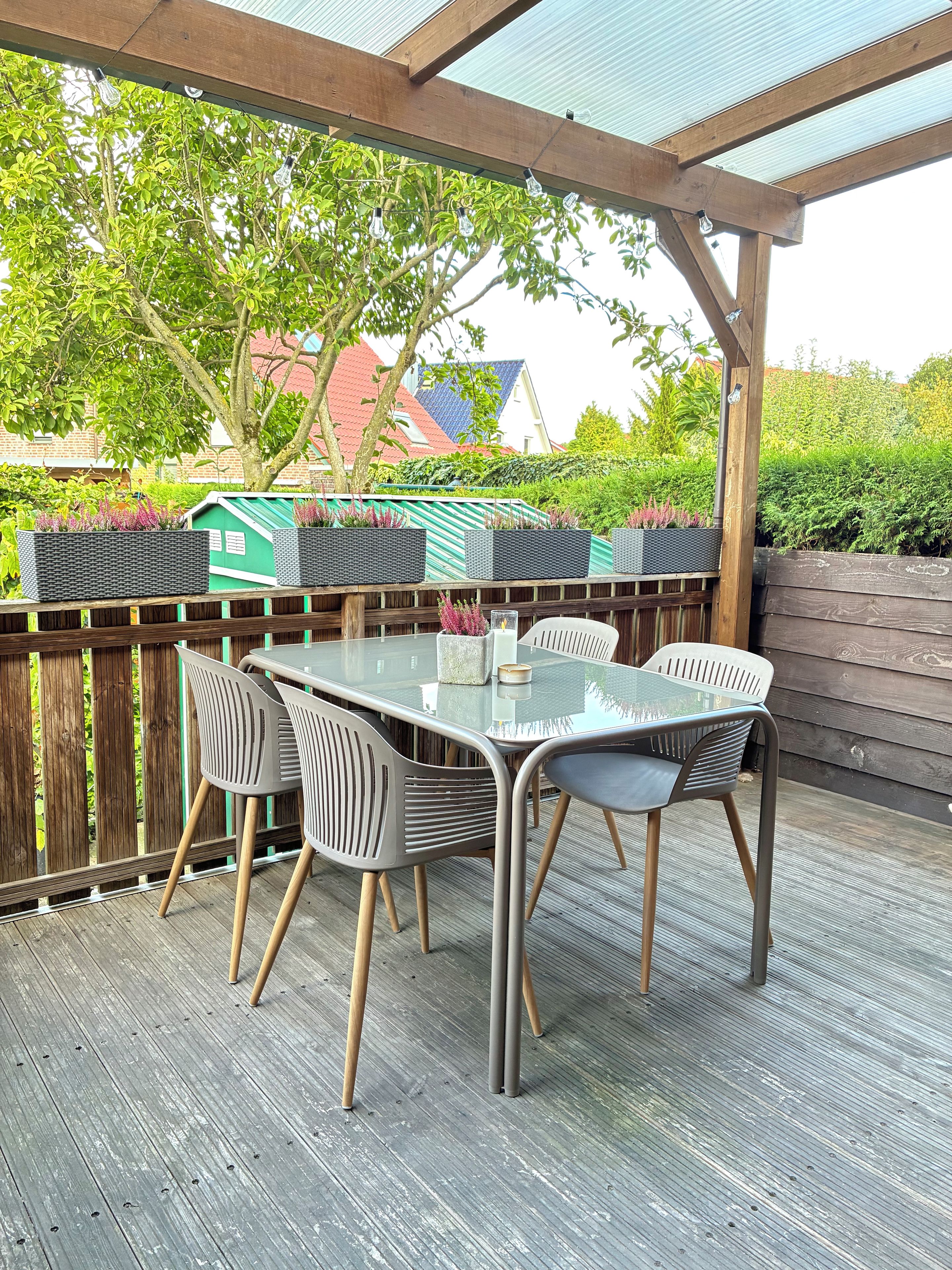 An outdoor patio with a glass-top table and chairs, surrounded by a wooden deck and a wooden fence.