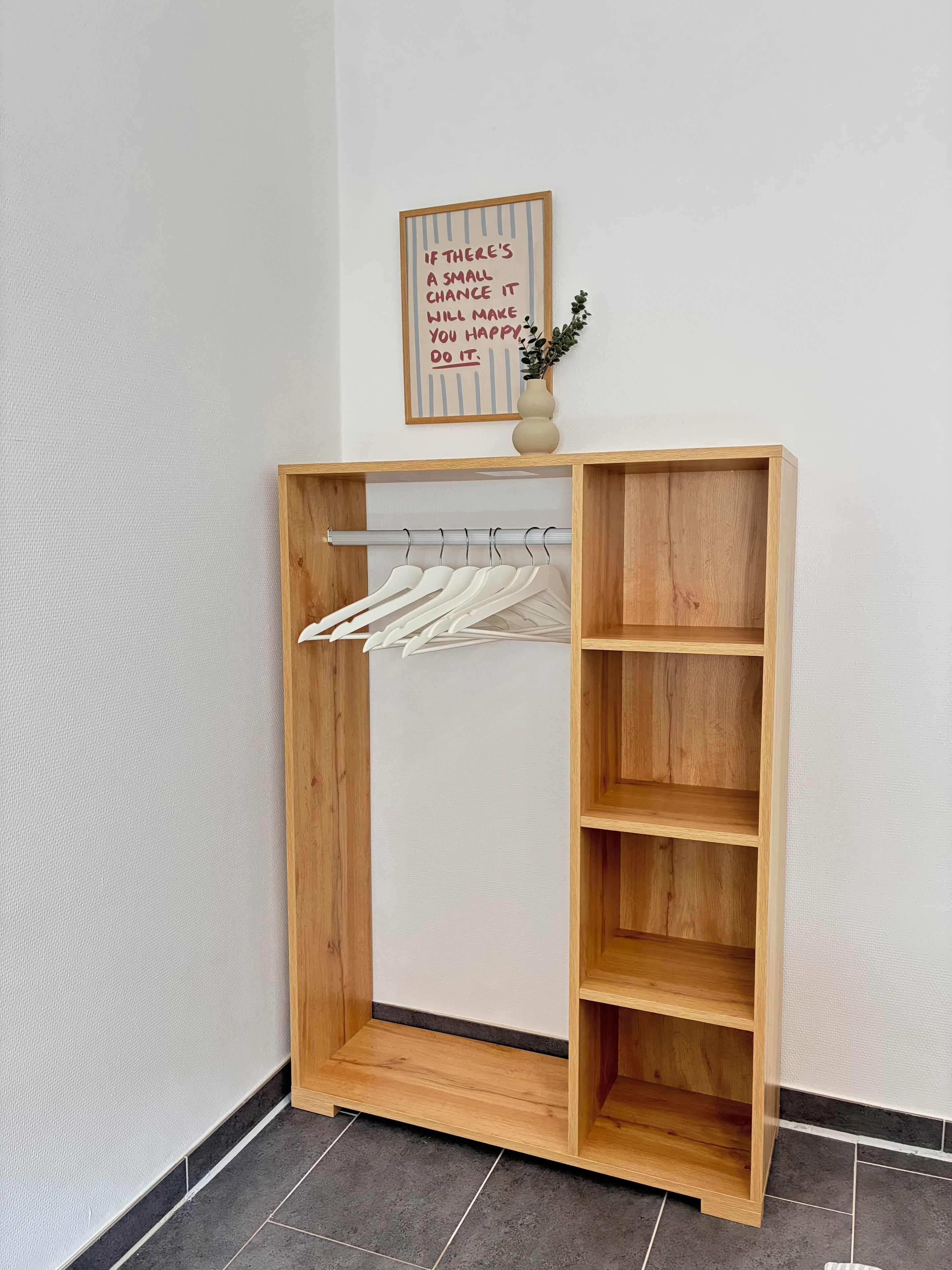 Ein Holzkleiderkasten mit Kleiderstange und Regalen, eine kleine Vase mit Pflanze und ein motivierendes Zitat an der Wand.