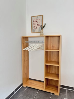 A wooden wardrobe with a hanging rack and shelves, a small vase with a plant, and a motivational quote on the wall.