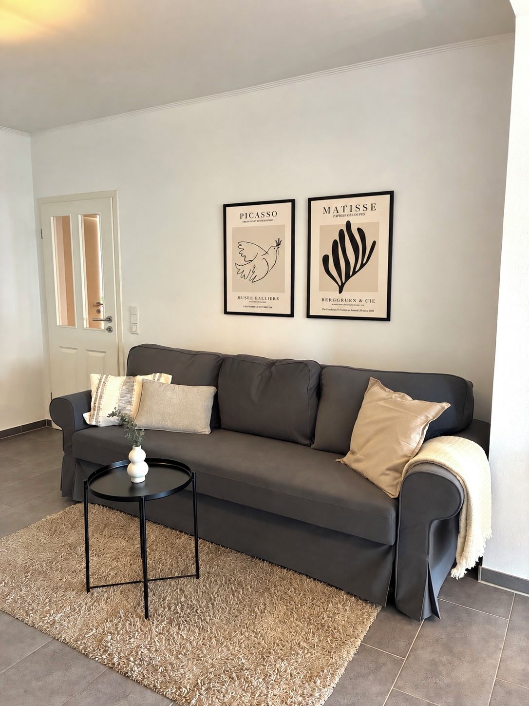 A living room with a dark grey sofa, a round black coffee table, and a beige shag rug. There are two framed artworks on the wall.