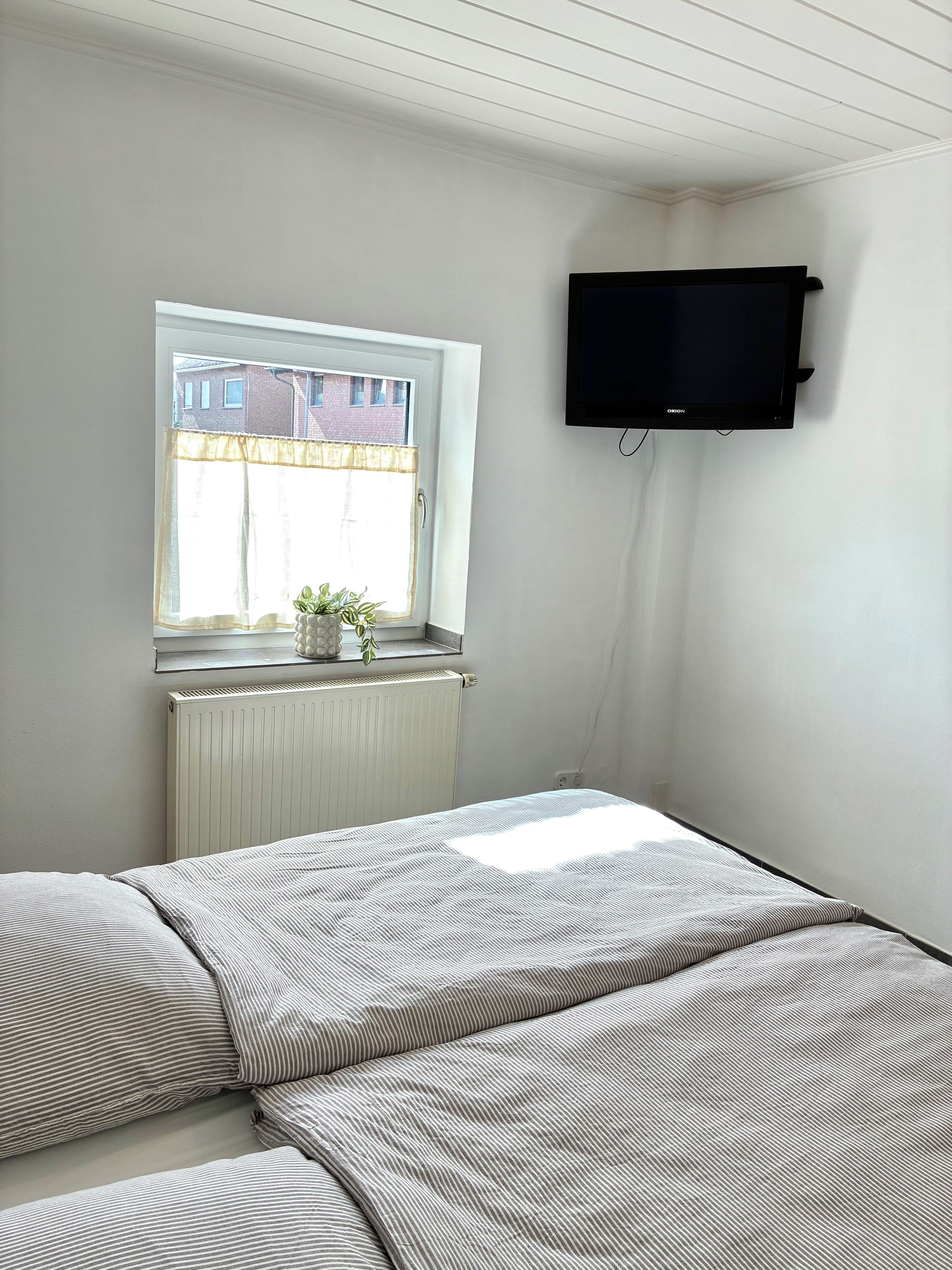 Un dormitorio con cama, ventana con vista a edificios, radiador y televisor montado.