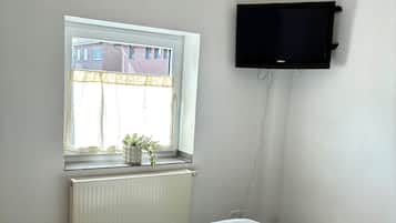 Un dormitorio con cama, ventana con vista a edificios, radiador y televisor montado.