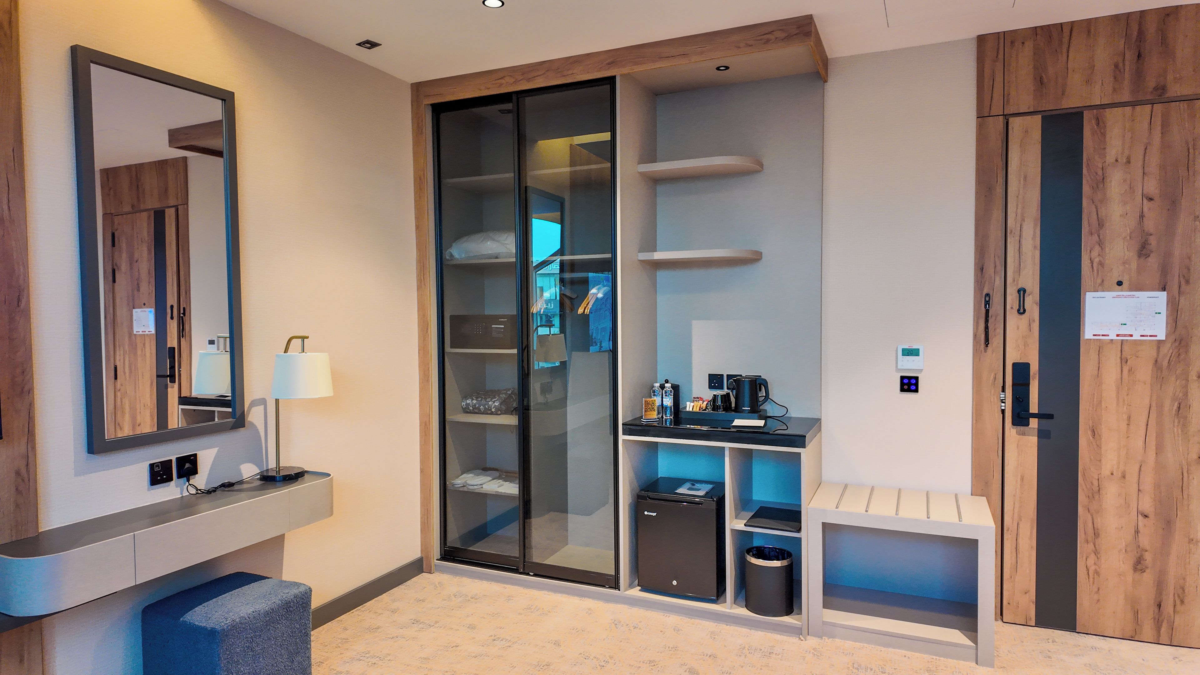 A modern hotel room with a large mirror, a desk with a lamp, a wardrobe, and a built-in wardrobe with shelves.