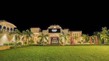 A large, well-lit building with a prominent entrance, surrounded by a manicured lawn and palm trees.