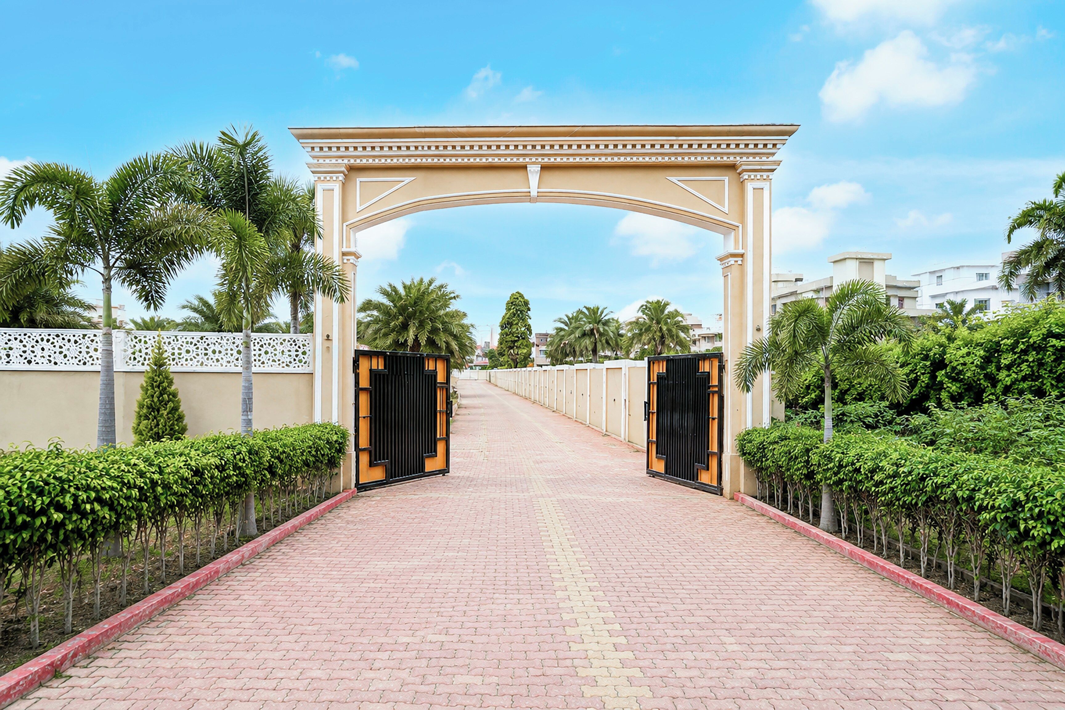 Un camino pavimentado que conduce a una entrada con puerta de hierro forjado y un arco decorativo, flanqueado por setos bien cuidados y palmeras.