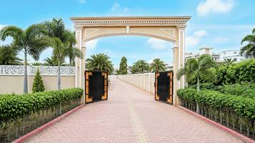 Un camino pavimentado que conduce a una entrada con puerta de hierro forjado y un arco decorativo, flanqueado por setos bien cuidados y palmeras.