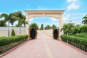 A paved pathway leading to a gated entrance with a decorative archway, flanked by neatly trimmed hedges and palm trees.
