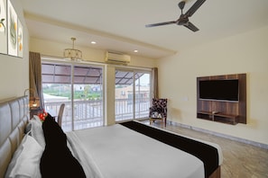 Una habitación de hotel con una cama, un televisor montado en la pared, un ventilador de techo y un balcón con vistas a la ciudad.