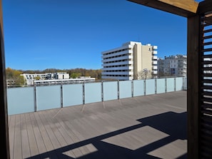 A wooden deck with a view of a multi-story building with a 'San' sign, a river, and trees in the background. - Elegant Vacation Rental “Apartamenty-plaza” – 300 m from the beach, Kolberg (Kołobrzeg)