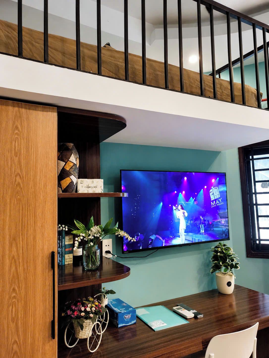 Una sala moderna con un televisor de pantalla plana montado en la pared, un mueble de madera con adornos, un escritorio con una planta y una escalera con barandilla de madera.