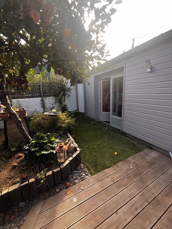 Un patio trasero con una terraza de madera, un pequeño jardín con plantas y un bebedero para pájaros, y una casa blanca con puerta corrediza de vidrio.