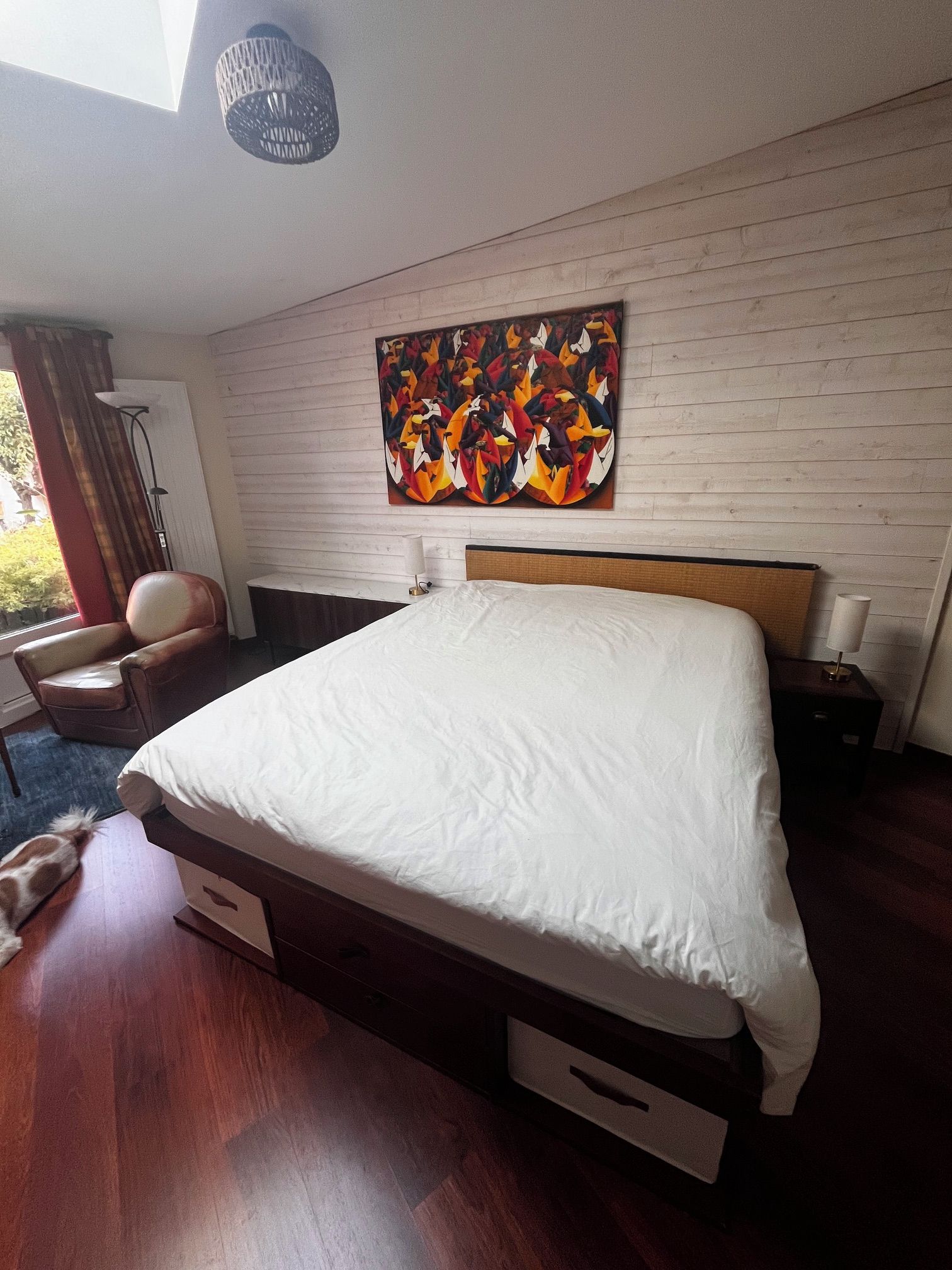 A modern hotel room with a large bed, a wooden floor, a wall-mounted painting, a chair, and a desk.