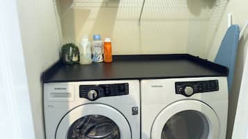 Dos lavadoras Samsung de carga frontal en un cuarto de lavado, con un estante superior que contiene varios objetos.