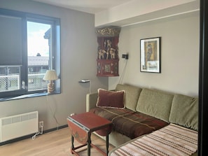 A living room with a sofa, a small table, a radiator, a window with blinds, a cabinet with figurines, and a framed picture on the wall. - Modern apartment with sauna by Sjusjøvannet (Sjusjøen)
