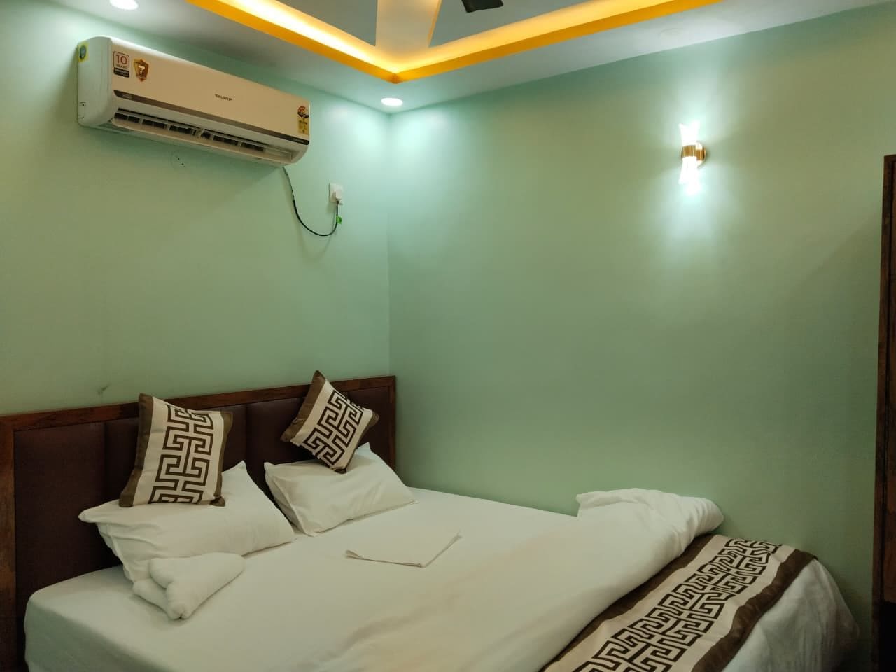 Een hotelkamer met een bed, kussens, een airconditioningapparaat en een wandlamp.