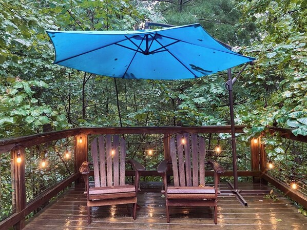 A wooden deck with two chairs and a blue umbrella, surrounded by greenery.
