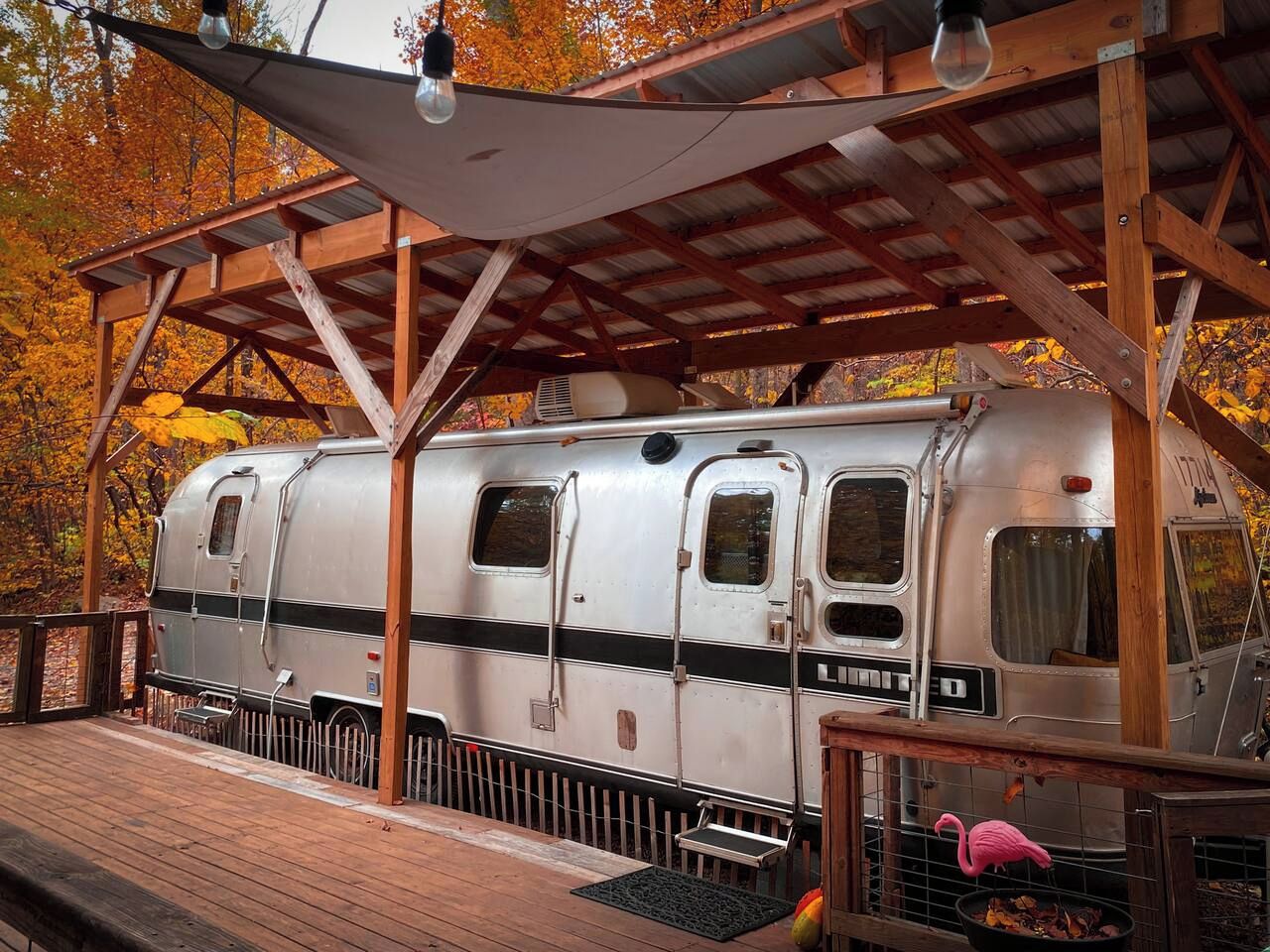 Une caravane ancienne est garée sous un pavillon en bois doté d’un toit en métal, et elle est entourée du feuillage automnal.