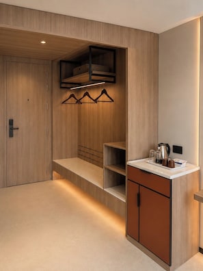 Una habitación de hotel moderna con un armario de madera, una estantería empotrada con ropa colgada y una pequeña kitchenette, además de una mesita de noche.