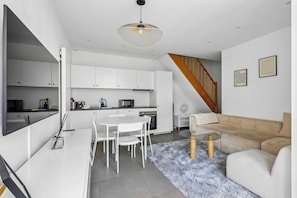 A modern kitchen with white cabinets, a dining table with chairs, a staircase with wooden steps, and a living area with a sofa and a glass coffee table. - Cozy 2-Bedroom House by Made For You (Neuilly-sur-Seine)