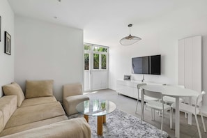 A modern living room with a glass coffee table, beige sofa, and a dining area with a white table and chairs. - Cozy 2-Bedroom House by Made For You (Neuilly-sur-Seine)