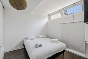 Un dormitorio con cama, almohadas, toallas enrolladas, una lámpara en el techo, una ventana y un televisor instalado en la pared.