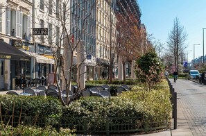 Eine Reihe von Sitzgelegenheiten im Freien mit Tischen und Stühlen, ein Bürgersteig und ein Gebäude mit mehreren Geschäften im Hintergrund.