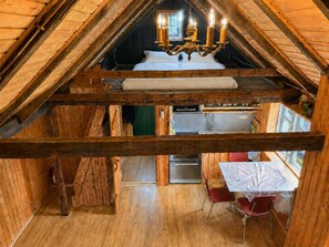 A loft-style room with a sloped ceiling, wooden beams, a chandelier, a bed, a dining table with chairs, and a kitchen area.