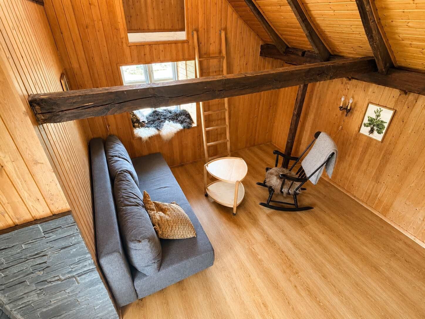 Een gezellige kamer met een houten plafond, een grijze bank, een schommelstoel, een klein tafeltje en een ladder.