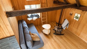 Een gezellige kamer met een houten plafond, een grijze bank, een schommelstoel, een klein tafeltje en een ladder.