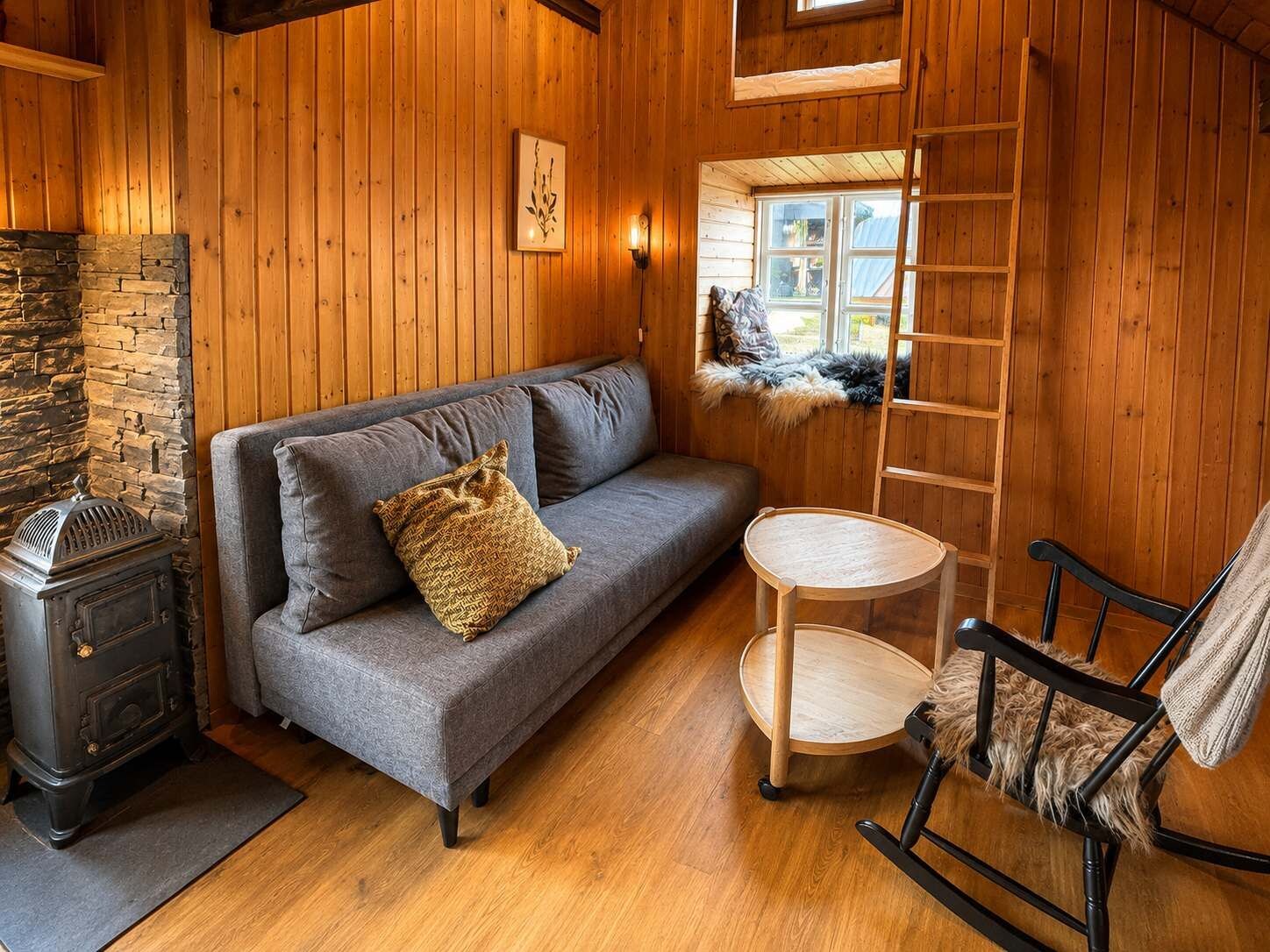 A cozy cabin interior with a stone fireplace, a sofa with a throw pillow, a wooden ladder, and a chair with a fur throw.