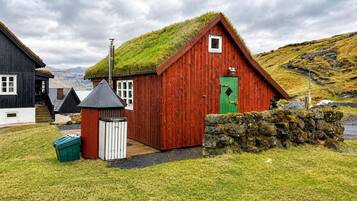 A traditional red wooden house with a grass roof, a stone wall, and a small shed.