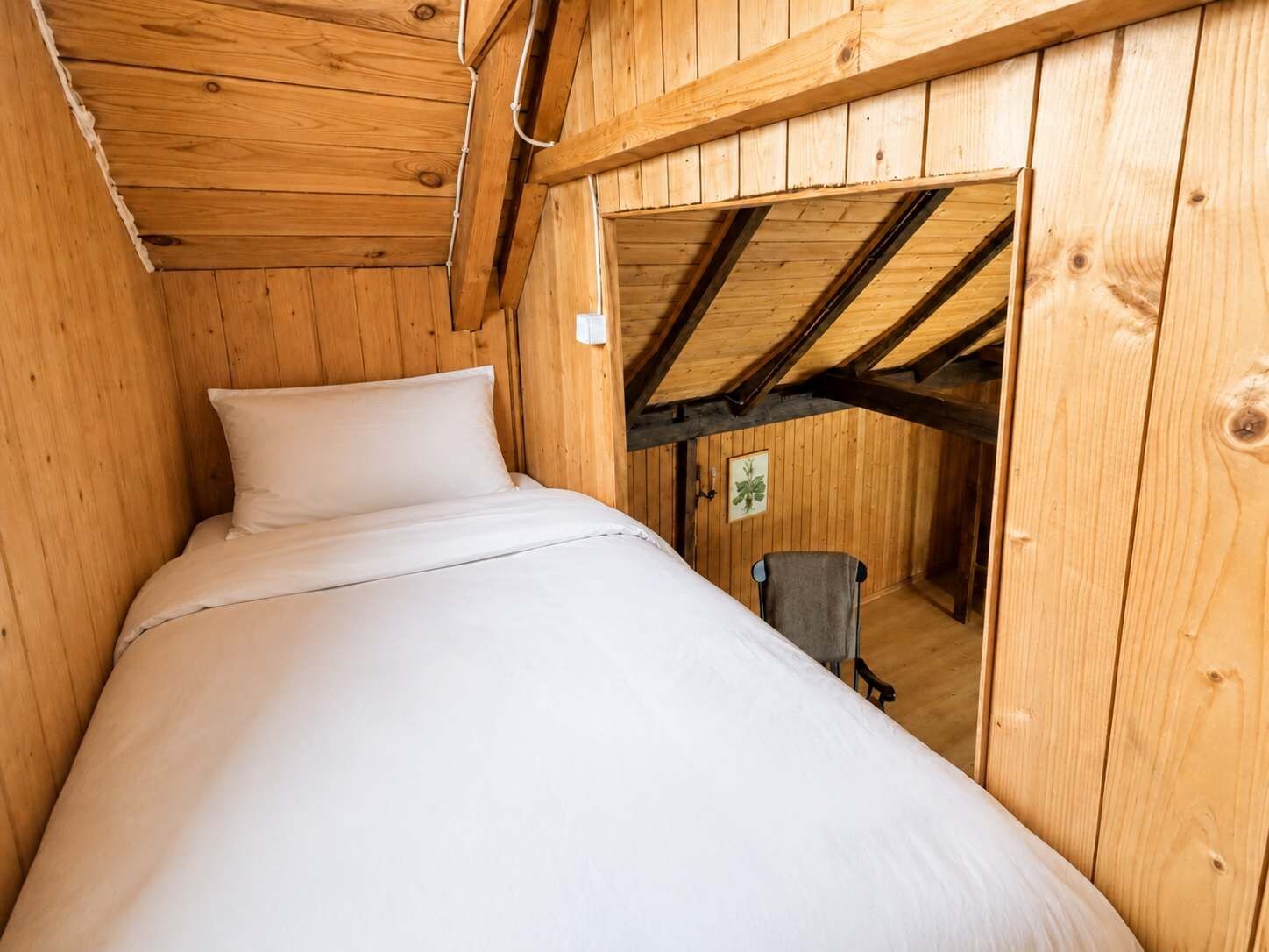 Ein Zimmer mit Holzboden, einem Bett, einem Sessel und einer Tür, die in einen anderen Raum führt.