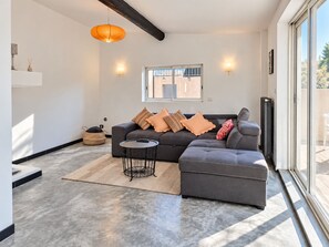 Un salon moderne avec un canapé d’angle, une table basse ronde et une grande fenêtre offrant une vue sur l’extérieur.