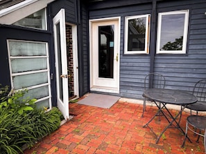 A house with a dark exterior, a front door, a small outdoor seating area with a table and chairs, and a brick pathway leading to the door.