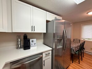 A compact kitchen with white cabinets, a stainless steel refrigerator, a coffee maker, and a dining area with a table and chairs. - Freshly Updated Creekside Condo Near Sugar Mountain Resort (Sugar Mountain)