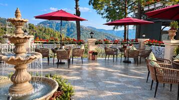 Un patio al aire libre con una fuente escalonada, muebles de mimbre, sombrillas rojas y montañas al fondo.