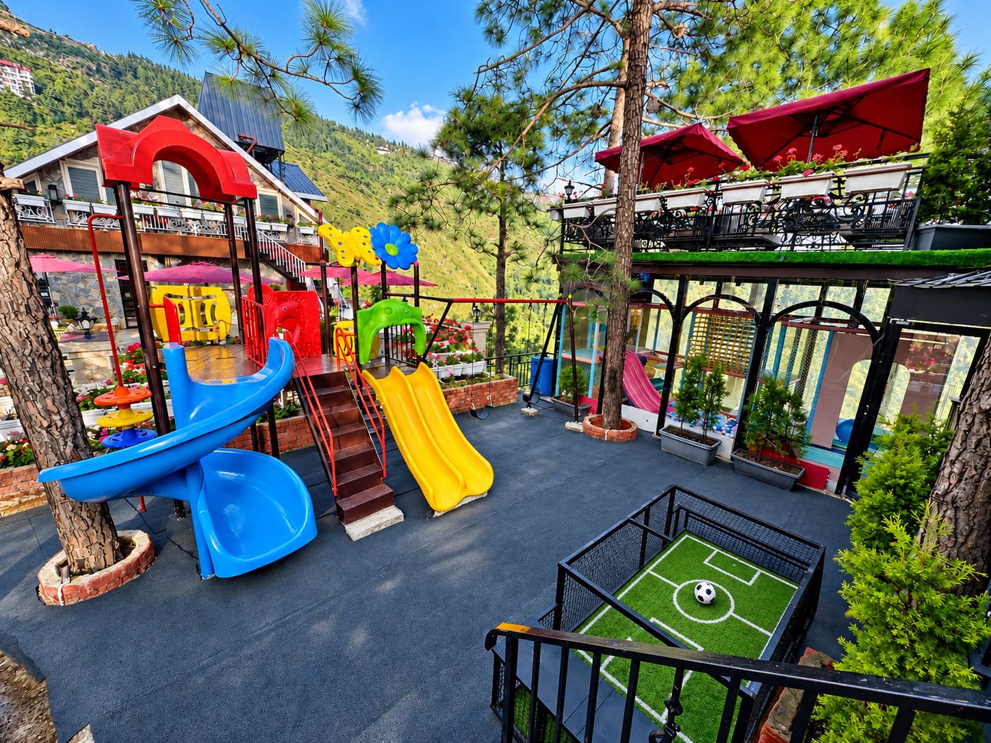 A playground with slides and climbing structures, surrounded by potted plants and a building with a red roof.